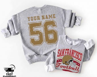 Op de afbeelding: Twee grijze sweatshirts. Het grotere sweatshirt heeft "YOUR NAME" en het nummer "56" in goud. Het kleinere sweatshirt heeft "SAN FRANCISCO Football" met een gouden en rode footballhelm.