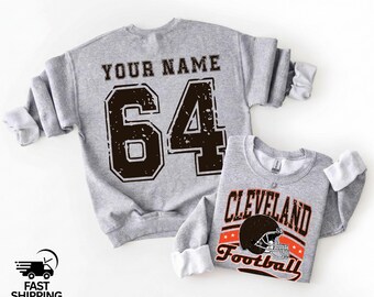 May include: Two gray sweatshirts. The larger one has the text "YOUR NAME" above the number "64" in a distressed font. The smaller sweatshirt says "CLEVELAND Football" with a football helmet graphic. Both have white cuffs.