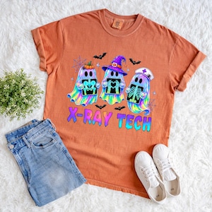 May include: A burnt orange short-sleeve t-shirt featuring a graphic of three iridescent ghosts holding X-ray images. One ghost wears a witch hat, another a nurse cap. The text X-RAY TECH is printed in blue and purple below the ghosts with bats.