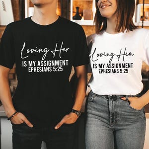 May include: Black and white matching t-shirts. The black shirt says "Loving Her is my assignment Ephesians 5:25" in white script. The white shirt says "Loving Him is my assignment Ephesians 5:25" in black script. The shirts are worn by a couple.