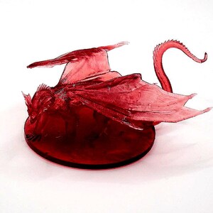 Könnte beinhalten: Eine durchscheinende rote Drachenfigur mit ausgebreiteten Flügeln und einem eingerollten Schwanz. Der Drache ist auf einer kreisförmigen Basis positioniert, mit komplizierten Details in seinen Flügeln und seinem Körper. Die Figur steht vor einem weißen Hintergrund.