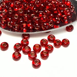 May include: A collection of small, round, transparent red glass beads with a metallic gold-colored hole. The beads are scattered on a white surface and piled in a silver spoon. These beads are ideal for jewelry making and crafts.