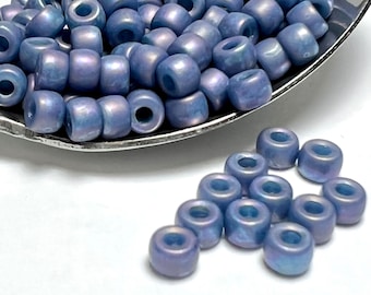 May include: A close-up of a pile of small, light blue glass beads with a iridescent finish. The beads are in a metal spoon and some are scattered on a white surface.