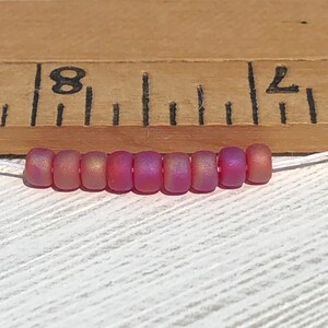 May include: A strand of eight iridescent pink seed beads, approximately 8 inches long, against a wooden ruler with markings for inches.