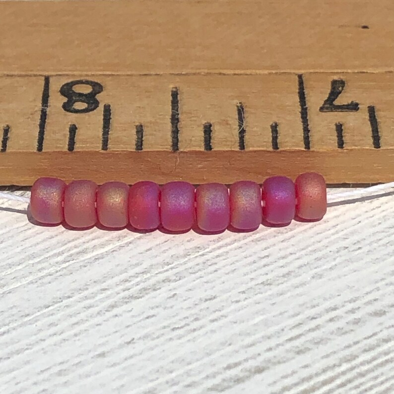 May include: A strand of eight iridescent pink seed beads, approximately 8 inches long, against a wooden ruler with markings for inches.