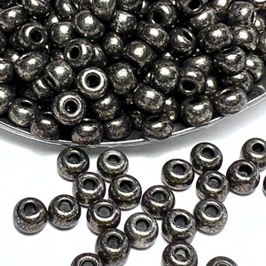 May include: A close-up of a pile of small, round, black beads with a metallic finish. The beads are scattered on a white surface.