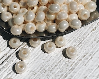 May include: A collection of small, round, off-white beads with a pearlescent, glossy finish. Each bead features a central hole for threading. The beads are clustered together in a metallic container, with several individual beads scattered nearby.