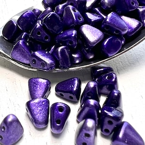 May include: A close-up shot showcases a collection of triangular, dark purple beads with two holes each. The beads are glossy and appear to be made of glass or a similar material. Some beads are in a silver spoon, while others are scattered around.