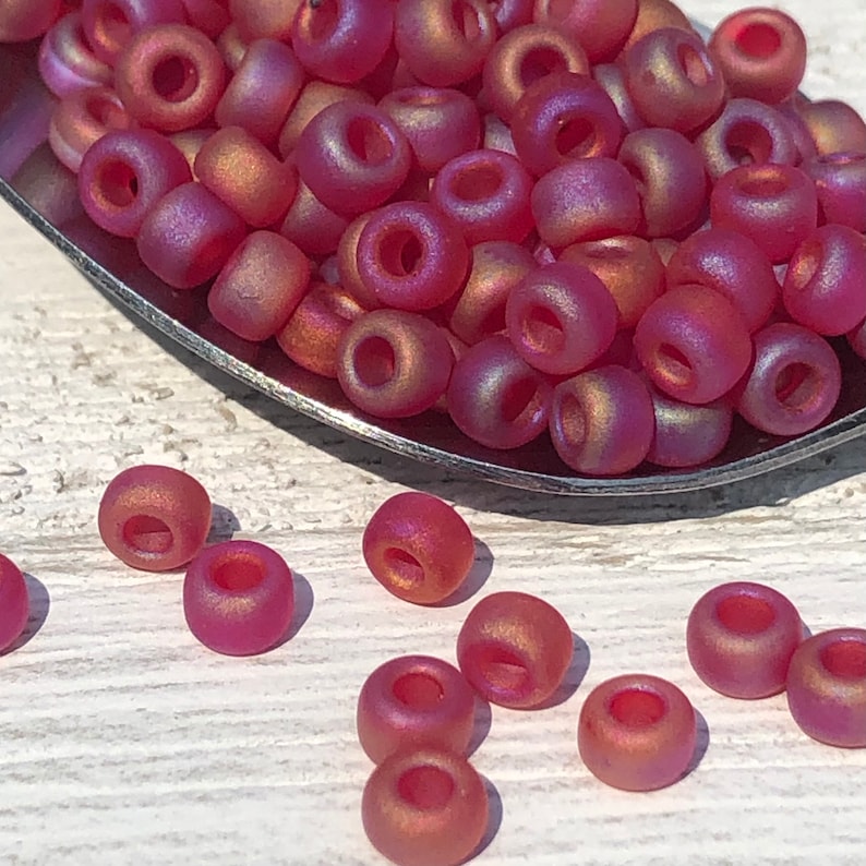 May include: A close-up of a pile of small, round, iridescent pink beads. The beads are in a metal spoon and some are scattered on a wooden surface.