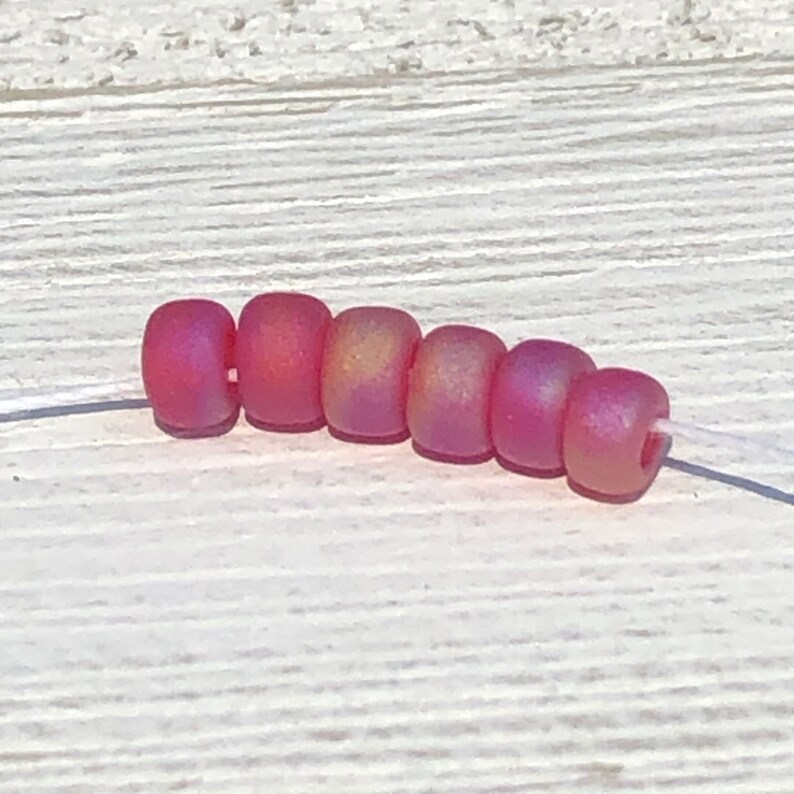 May include: A strand of eight small, round, pink glass beads with a slight iridescent shimmer. The beads are strung on a white thread.