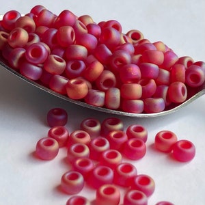 May include: A close-up of a pile of small, round, iridescent pink beads. The beads are arranged in a pile on a silver spoon. There are also some beads scattered around the spoon.