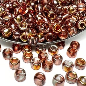 May include: A collection of small, metallic beads in shades of copper, bronze, and gold. The beads are cylindrical with rounded edges and a reflective, glossy finish. Some beads are scattered on a white surface.