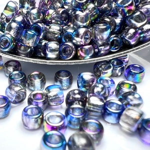 May include: A close-up of iridescent blue glass beads, some in a silver spoon and others scattered on a white surface.