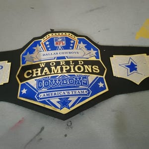 May include: A black and gold championship belt with blue accents. The belt features the text "WORLD CHAMPIONS COWBOYS" and "AMERICA'S TEAM". The belt also has the NFL logo and star designs. The words "DEFEND THE STAR" are also visible.
