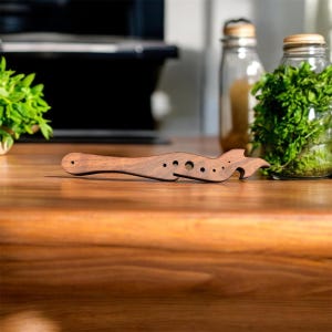 Puede incluir: Un abridor de botellas y utensilio de cocina de madera de color marrón oscuro. La herramienta tiene una serie de agujeros y un diseño curvo, con un abridor de botellas en un extremo. Está sobre una superficie de madera.