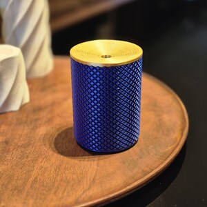 May include: A cylindrical object featuring a vibrant blue body with a textured diamond-patterned surface and a flat, circular gold-coloured top with a central hole and concentric ring details. The item stands upright with a smooth, solid base.