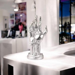 Könnte beinhalten: Eine silberfarbene Roboterhand-Skulptur. Die Hand, in einer Geste geformt, zeigt detaillierte mechanische Elemente. Die Skulptur steht auf einem kreisförmigen Sockel und einer weißen Oberfläche.