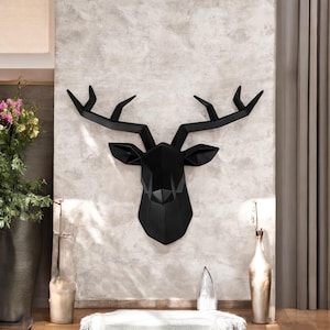 Könnte beinhalten: Mattschwarzer, geometrischer Hirschkopf als Wanddekoration. Der Hirschkopf ist an einer hellgrauen, strukturierten Wand befestigt. Das Geweih ist lang und kantig. Modernes, minimalistisches Design.