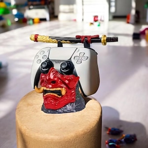 Könnte beinhalten: Ein weiß-schwarzer Gaming-Controller mit roter Oni-Maskenbasis und dekorativem Katana-Schwert. Der Controller hat schwarze Knöpfe und einen weißen Körper. Das Katana ist schwarz mit goldenen Akzenten. Die Maske ist rot mit goldenen Stoßzähnen.