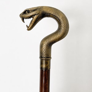 May include: A decorative walking cane with a snake head handle. The snake head is bronze-colored with detailed scales and open fangs. The shaft is a dark brown wood, and the cane has a brass-colored collar.