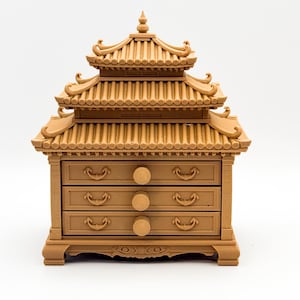 Peut inclure: Une petite boîte à bijoux de couleur beige avec un toit de style pagode et trois tiroirs. La boîte présente un design détaillé avec des bords incurvés et de petites poignées sur chaque tiroir. L'esthétique générale rappelle l'architecture asiatique.