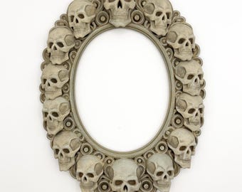 May include: Oval-shaped decorative frame with a gray finish, adorned with numerous skull embellishments. The frame's design features a repeating pattern of skulls around the oval opening, creating a gothic or Halloween theme.