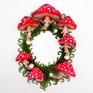 May include: A decorative wreath featuring red mushrooms with white spots, arranged around a green leafy base. The mushrooms have tan stems and are accented with small white pearls. The wreath is set against a white background.