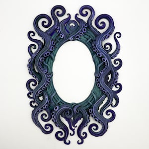 Può includere: Una cornice ovale con un interno verde acqua scuro e un bordo di tentacoli di polpo blu scuro stilizzati. I tentacoli si avvolgono attorno alla cornice, creando un design unico e artistico. La cornice è montata su uno sfondo bianco.