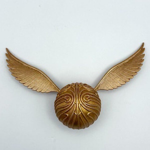 Peut inclure: Un vif-or doré, une balle fictive de la série Harry Potter, est présenté sur un fond blanc. La balle a une surface texturée avec des détails complexes et deux ailes déployées. Les ailes sont également dorées et ont un aspect de plumes.