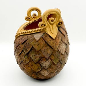 May include: A decorative, egg-shaped container with a textured, scale-like pattern in shades of brown and green. The top features a gold-colored, ornate design. The interior is a deep red. This unique piece could be used as a decorative item or a small container.