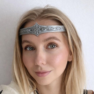 May include: A silver-colored metal headband with an ornate design. The headband is worn on the forehead. The person in the image is wearing a cream-colored top with floral embroidery.