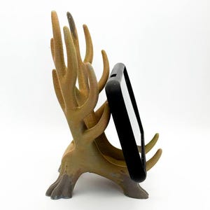 Puede incluir: Un soporte para teléfono en forma de árbol marrón con una funda negra para teléfono. El soporte tiene múltiples ramas y una superficie texturizada, lo que proporciona una forma única y decorativa de sostener un teléfono. El soporte está hecho de un material duradero.