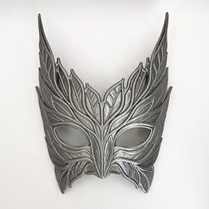 May include: A silver-colored masquerade mask with a leaf design. The mask has eye openings and is decorated with detailed leaf patterns. The mask has pointed extensions on the top, giving it a unique appearance.