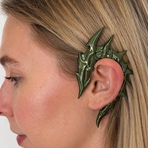 May include: A dark green ear cuff with a spiky, ornate design. The ear cuff is worn on the ear and has a metallic sheen. The design features sharp, pointed elements, creating a bold and edgy aesthetic. The ear cuff is a unique piece of jewelry.