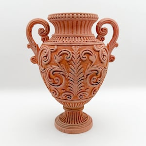 May include: A terracotta-colored decorative vase with intricate scrollwork and two curved handles. The vase features a detailed floral design and a fluted neck and base, showcasing a classical aesthetic.