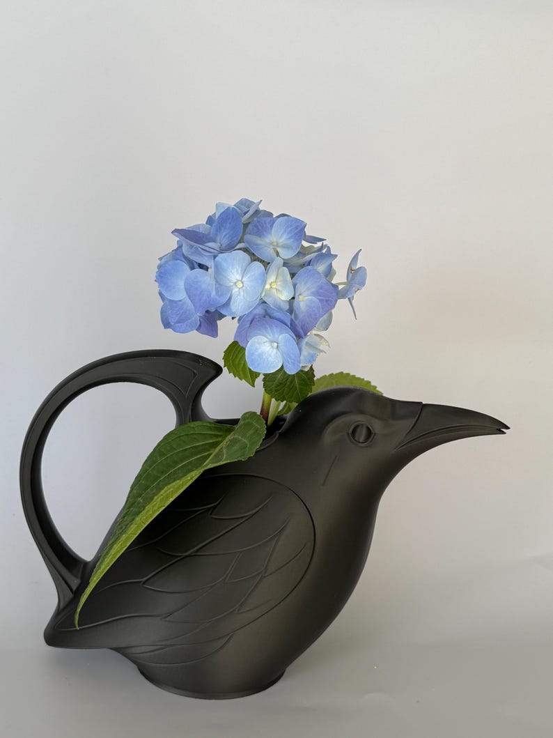 May include: A matte black bird-shaped watering can featuring a curved handle and a pointed beak. A cluster of vibrant blue hydrangea flowers with green leaves is placed inside the opening of the bird, extending upwards from the top of the vessel.