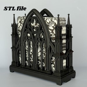 May include: A black napkin holder featuring an intricate architectural design with pointed arches, spires, and circular tracery. The holder contains a stack of napkins with a black and white floral pattern. The text STL file is visible in the corner.