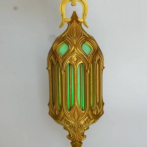 May include: A gold-colored lantern with an intricate, ornate frame featuring vertical bars and pointed arches. The interior of the lantern glows with a bright, solid green light. The structure includes a decorative top loop for hanging.