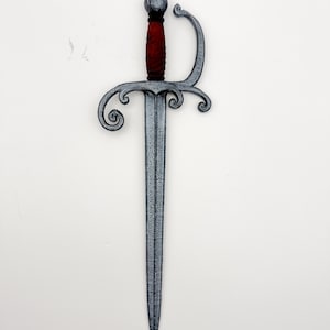 May include: A decorative sword with a silver-colored blade and guard, and a red handle. The guard features ornate, swirling designs. The sword is displayed against a plain white background, highlighting its detailed craftsmanship and historical aesthetic.