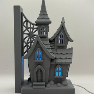 Könnte beinhalten: Graues, 3D-gedrucktes Geisterhaus-Nachtlicht mit blau leuchtenden Fenstern. Das Haus hat einen hohen, spitzen Turm und ein dekoratives Brückenelement. Das Design ist detailliert und skurril, perfekt für Halloween oder gruselige Dekoration.