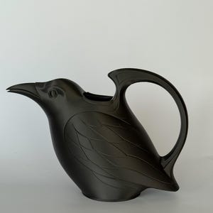 May include: A matte black watering can shaped like a bird. The vessel features a pointed beak spout, a curved handle extending from the back, and molded feather details along the body. The design is solid black with a smooth, uniform finish.