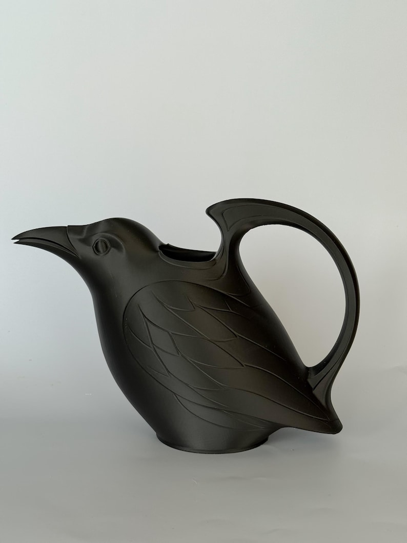 May include: A matte black watering can shaped like a bird. The vessel features a pointed beak spout, a curved handle extending from the back, and molded feather details along the body. The design is solid black with a smooth, uniform finish.