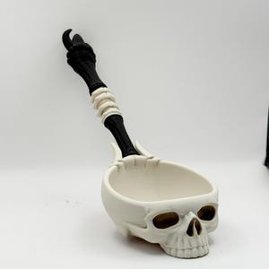 May include: A white skull-shaped serving spoon with a black handle and bone-colored accents. The handle features a detailed design, and the spoon's bowl is shaped like a skull. A unique kitchen utensil.