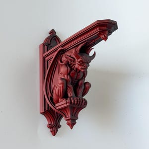 May include: A deep red wall-mounted corbel featuring a sculpted gargoyle figure with horns, wings, and muscular definition. The architectural support bracket has a pointed arch backplate and a flat top surface, finished in a matte red tone.