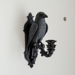 May include: A matte black wall-mounted candle holder featuring a sculpted raven perched on an ornate, scroll-detailed backplate. The bird faces right, with a decorative candle cup extending from the base, all finished in a uniform dark tone.