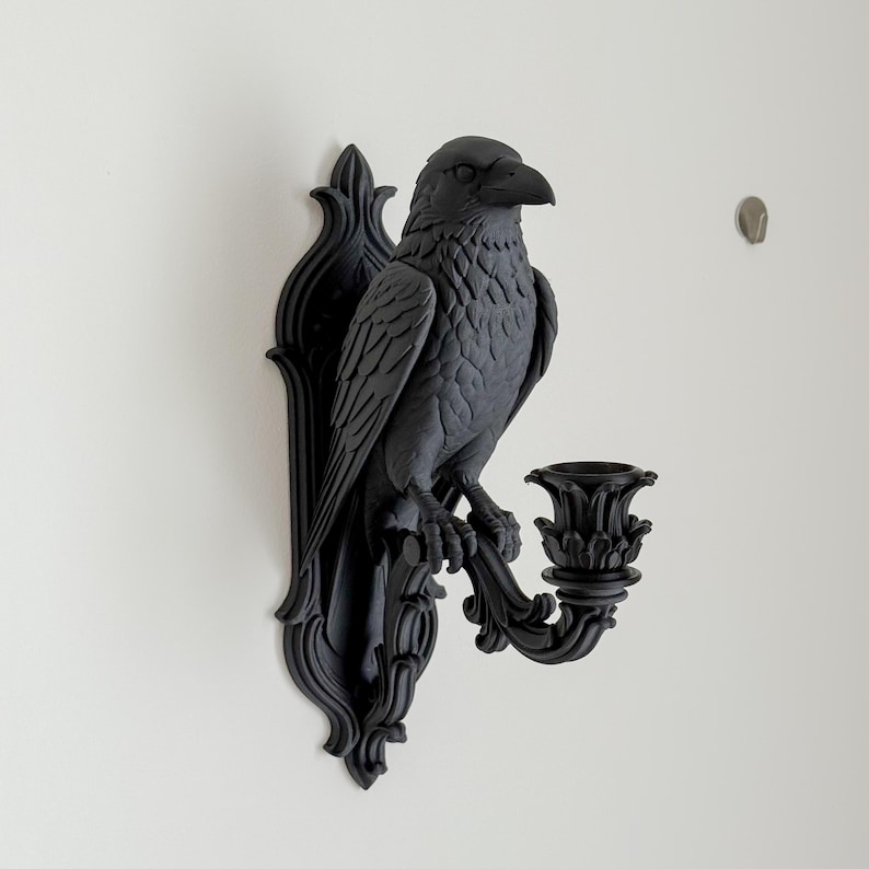 May include: A matte black wall-mounted candle holder featuring a sculpted raven perched on an ornate, scroll-detailed backplate. The bird faces right, with a decorative candle cup extending from the base, all finished in a uniform dark tone.