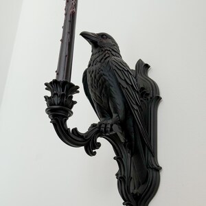 May include: A black wall-mounted candle holder featuring a sculpted raven perched on a decorative bracket. The raven holds a tall, slender black taper candle with visible wax drips and a small, glowing flame at the top, set against a plain wall.