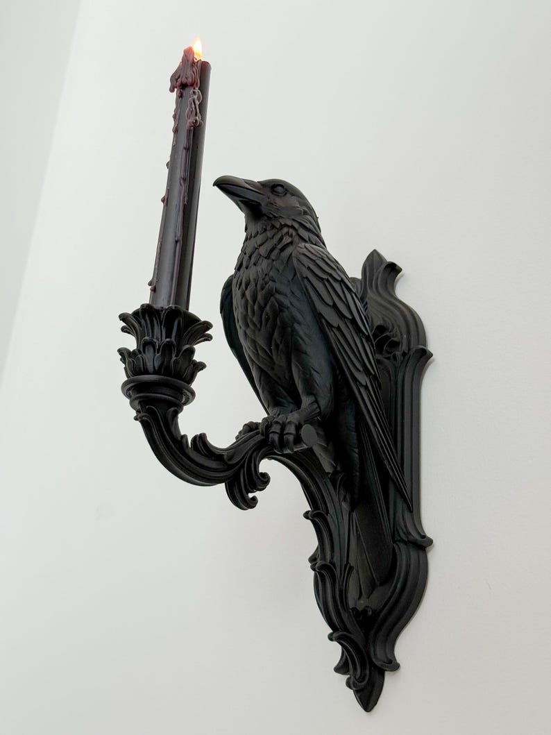 May include: A black wall-mounted candle holder featuring a sculpted raven perched on a decorative bracket. The raven holds a tall, slender black taper candle with visible wax drips and a small, glowing flame at the top, set against a plain wall.