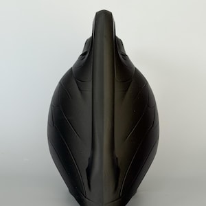 May include: A matte black sculptural object with a pointed, vertical central ridge and symmetrical, leaf-like embossed patterns curving outward along its sides. The form is elongated and tapered, featuring a smooth, dark surface texture throughout.