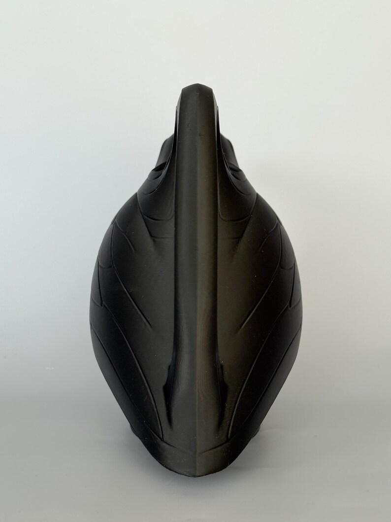 May include: A matte black sculptural object with a pointed, vertical central ridge and symmetrical, leaf-like embossed patterns curving outward along its sides. The form is elongated and tapered, featuring a smooth, dark surface texture throughout.